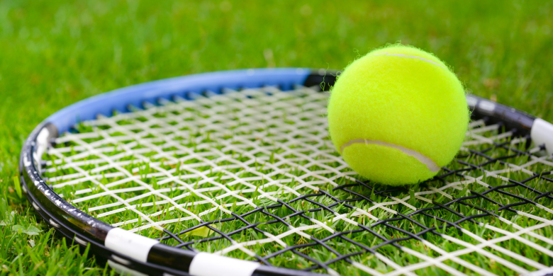 A tennis ball sits on top of a tennis racket on the grass.